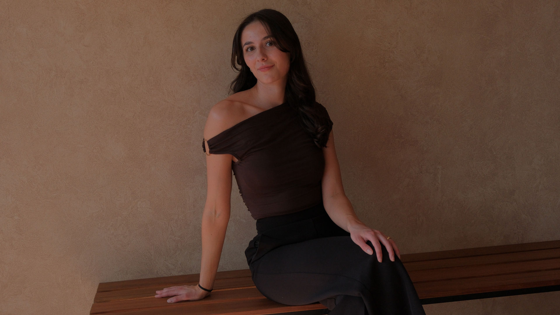 Emily Witt seated on a wooden bench, warm textured wall behind